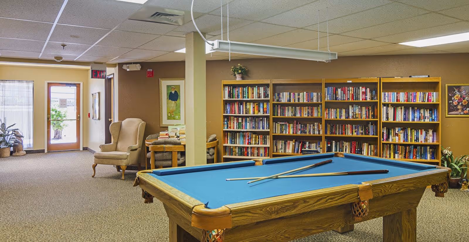 A cozy common area featuring a pool table with two cues resting on it, a large bookshelf filled with books, a comfortable armchair, a small round table with chairs, and framed artwork on the walls. The room has carpeted flooring and a drop ceiling with fluorescent lighting.