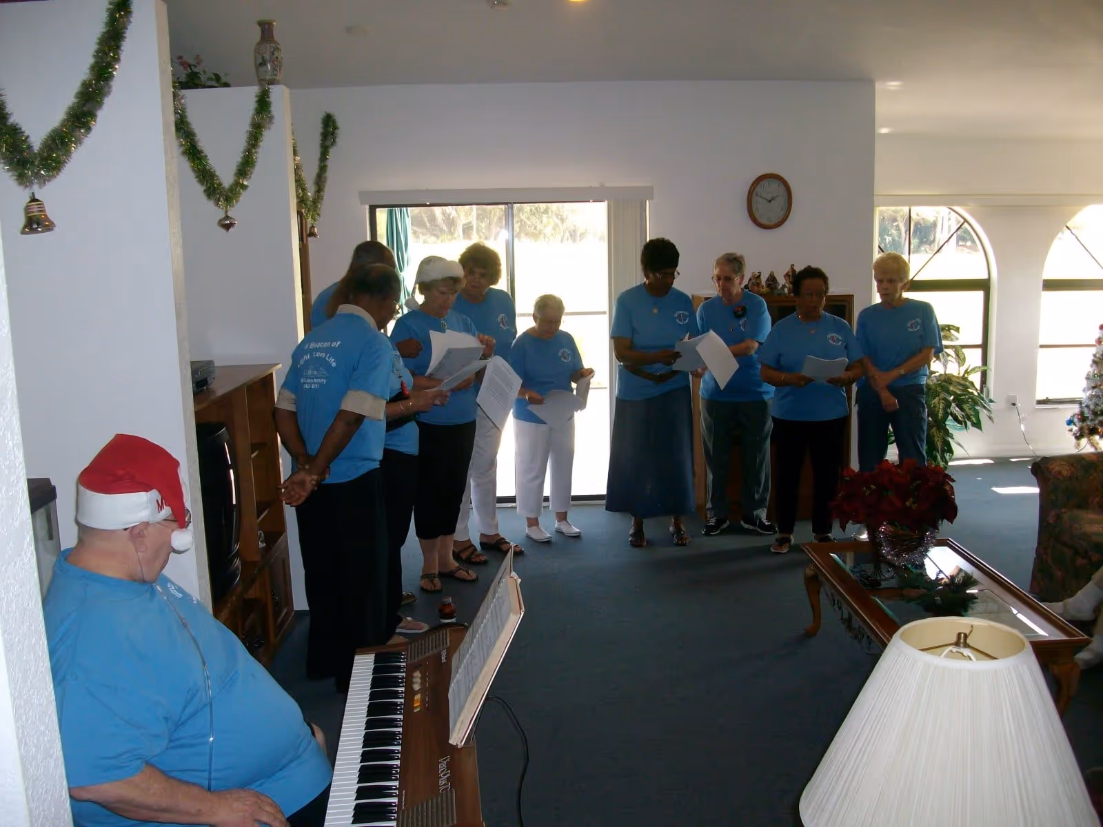 A group of elderly people wearing matching blue shirts are standing in a living room, holding papers and singing. One man is seated at a keyboard wearing a Santa hat. The room is decorated with Christmas garlands and a Christmas tree is visible in the background.