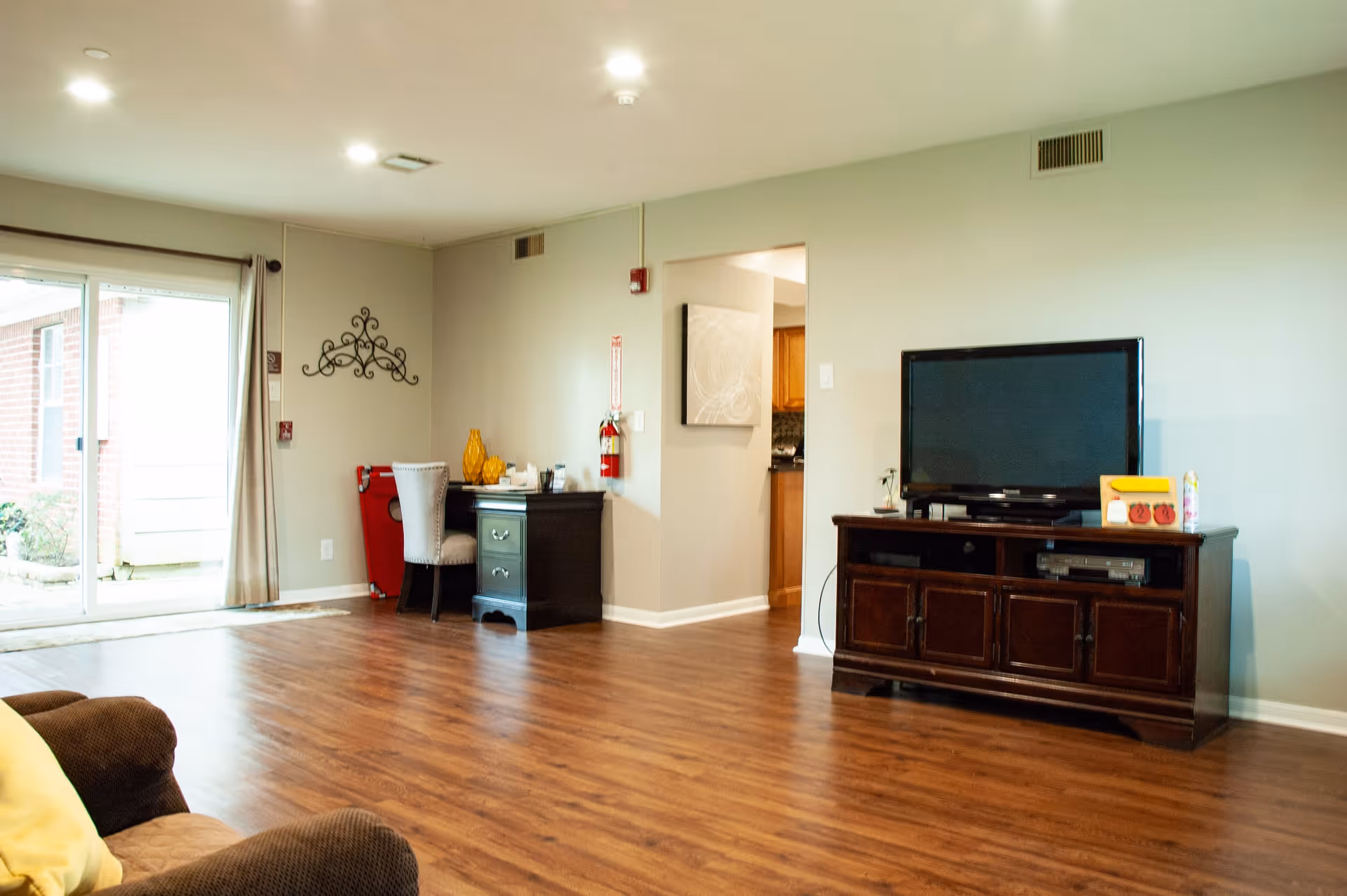 A spacious living room with wooden flooring, a brown couch with a yellow pillow, a dark wooden TV stand with a flat-screen TV, and a small desk with a chair and decorative items. There is a sliding glass door with curtains leading outside, and a fire extinguisher mounted on the wall near an open doorway to the kitchen.