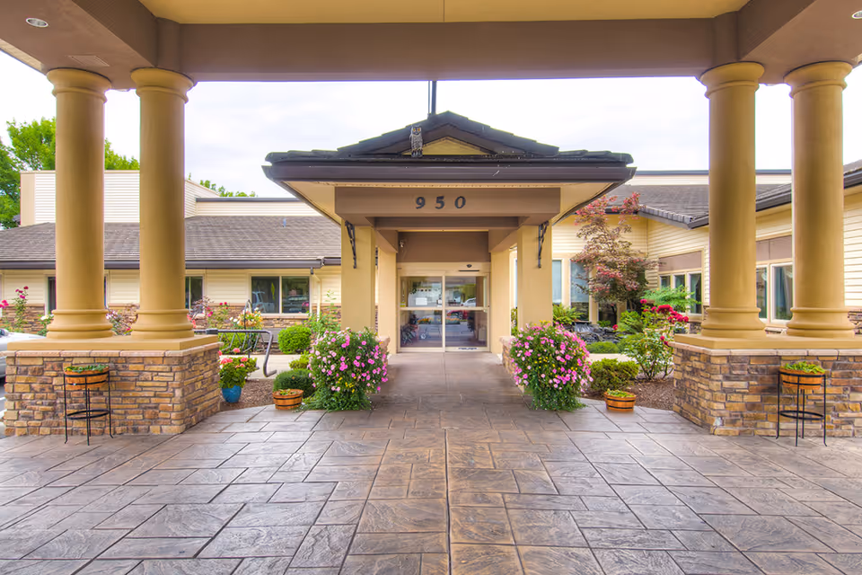 Entrance of Cascade Park Retirement Community featuring a covered driveway with large columns, potted plants with flowers, and a building with the number 950 above the doorway.