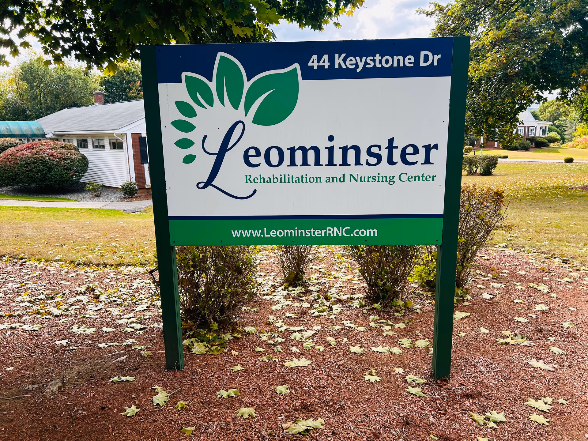 Outdoor sign for Leominster Rehabilitation and Nursing Center located at 44 Keystone Dr, with a green and white design featuring a leaf logo, surrounded by trees and bushes in a landscaped area.