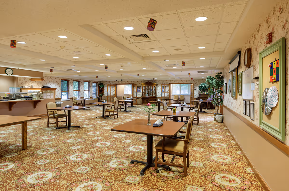 A spacious dining room with multiple wooden tables and chairs arranged neatly on a patterned carpet. The room has large windows along one wall letting in natural light, floral wallpaper, and ceiling lights. There are decorative plants and framed pictures on the walls, and a cabinet with glass doors at the far end. A counter area with stacked plates and cups is visible on the left side.