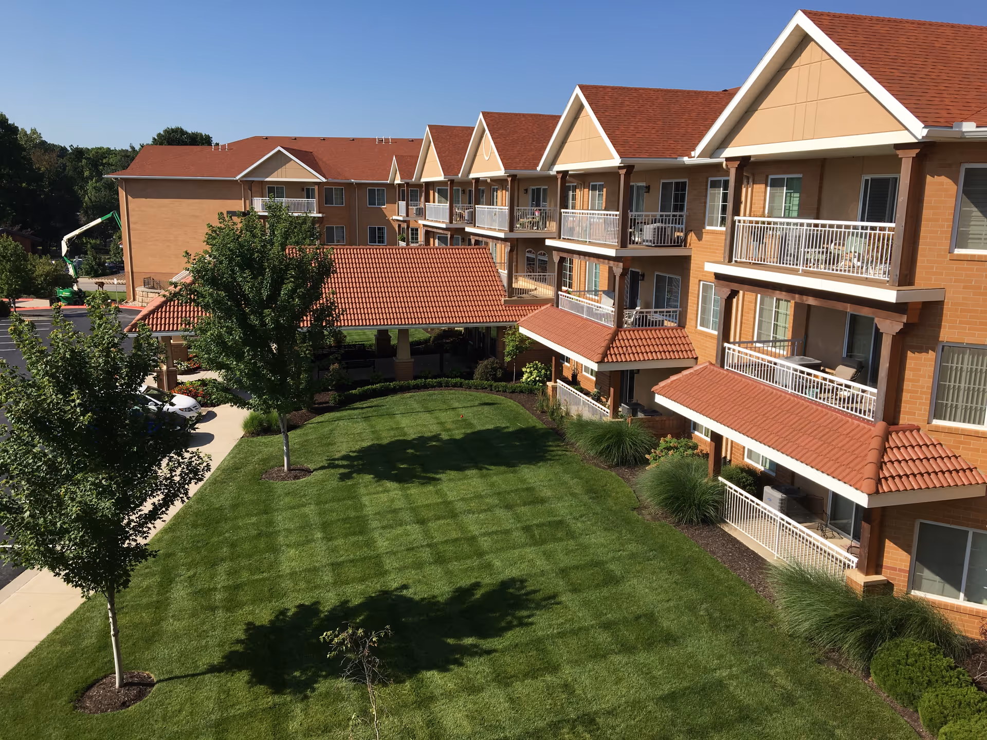 Exterior view of a senior living facility with a well-maintained green lawn, several trees, and a multi-story building with balconies and red-tiled roofs under a clear blue sky.