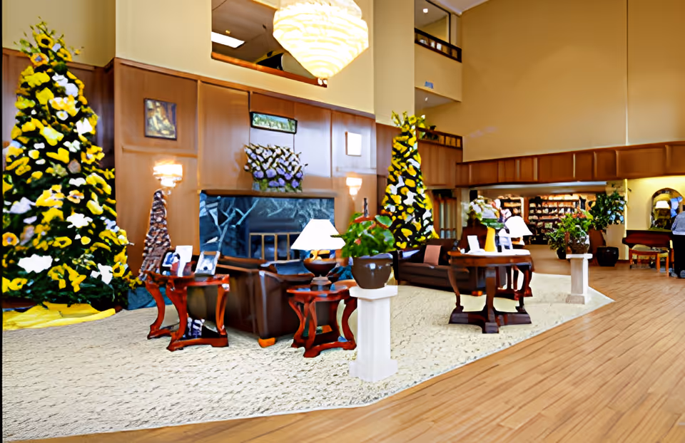 A spacious and well-lit living room area in a senior living facility with high ceilings, wooden paneling, and a large chandelier. The room features comfortable seating with sofas and chairs arranged around tables with lamps and potted plants. There are two large decorative Christmas trees adorned with yellow and white flowers. In the background, there is a fireplace and a bookshelf filled with books.