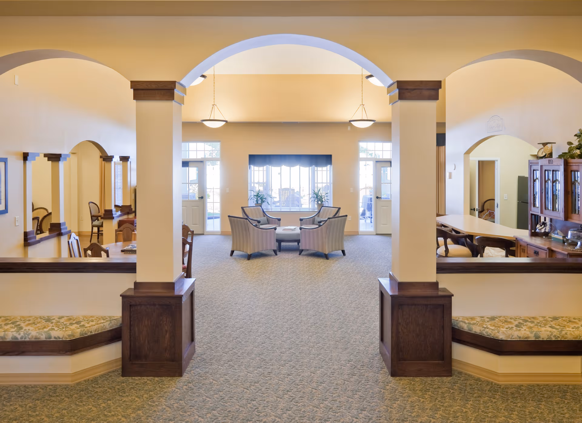 A spacious, well-lit common area in a senior living facility with beige walls and carpeted floors. The room features large archways with dark wood trim, built-in cushioned benches, and a seating area with four armchairs arranged around a small table near large windows and glass doors leading outside. There are pendant lights hanging from the ceiling and wooden furniture including a cabinet and a counter with chairs on the right side.