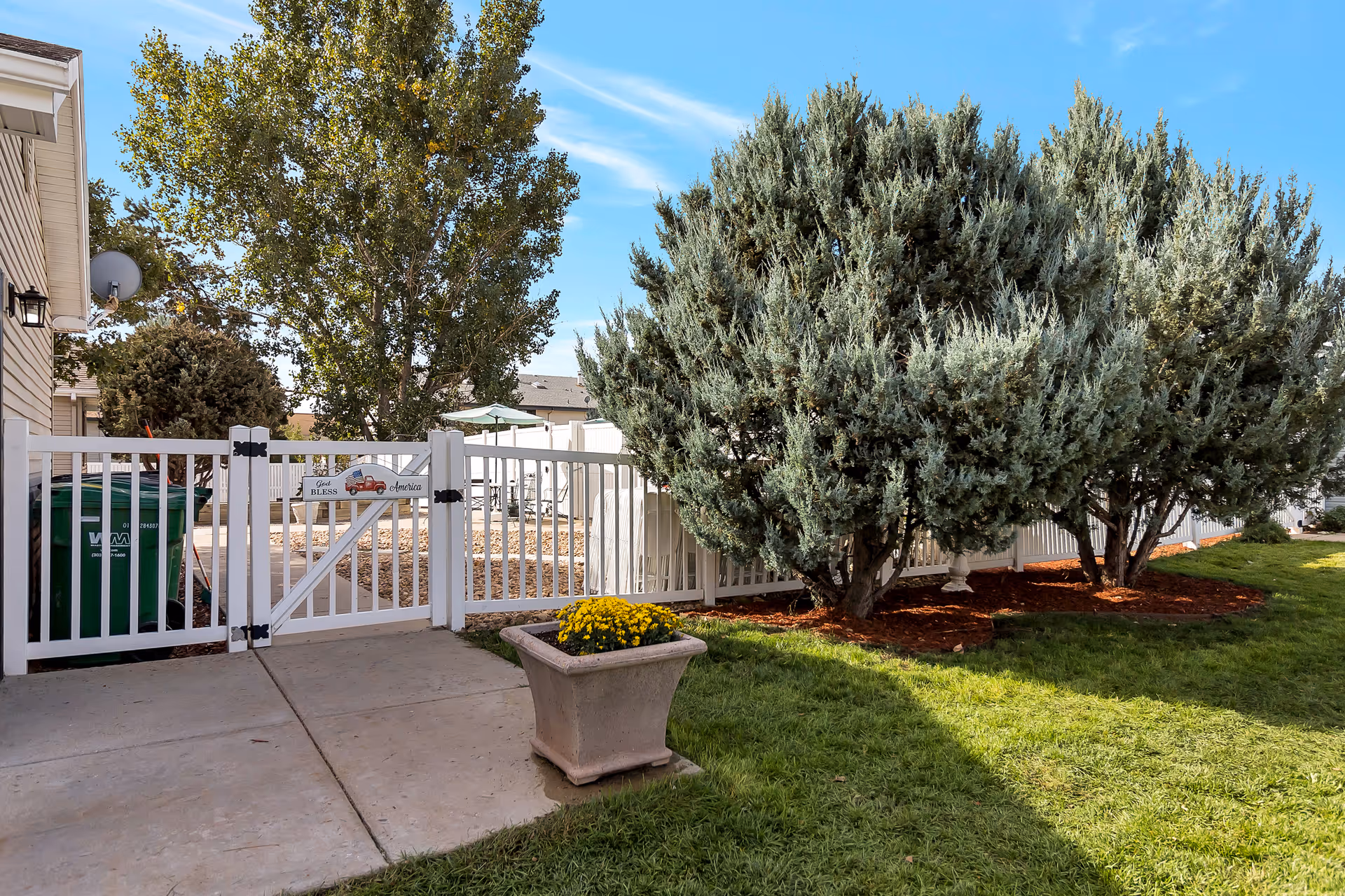 Outdoor view of a fenced garden area with a white gate and a sign that reads 'God Bless America'. There are green bushes, a tree, a concrete walkway, and a planter with yellow flowers. The sky is clear and blue.