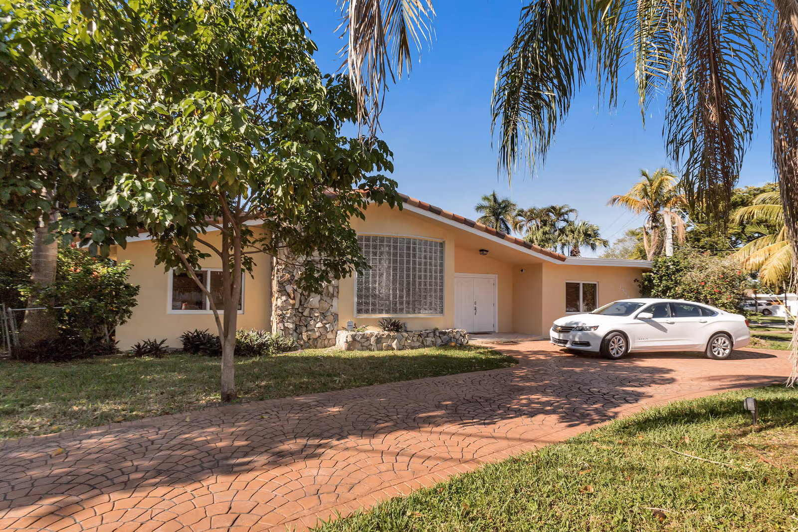 Front exterior view of a single-story assisted living facility building with a curved driveway, a white car parked on the driveway, palm trees, and other greenery under a clear blue sky.