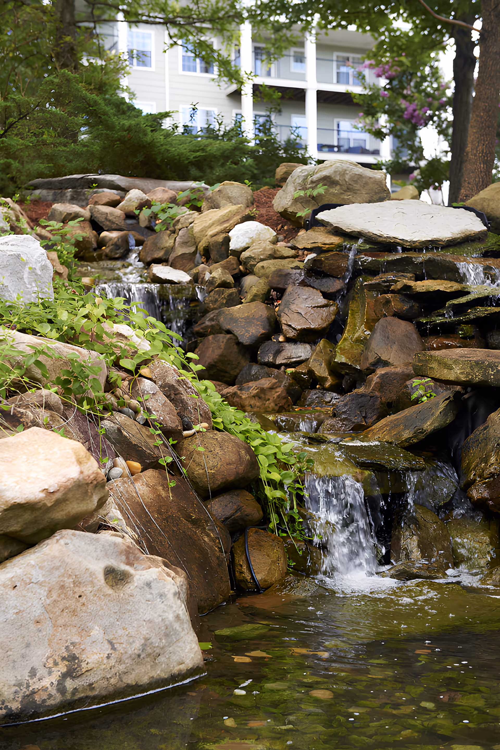 A landscaped outdoor area featuring a small cascading waterfall flowing over rocks into a pond, surrounded by greenery and plants. In the background, there is a multi-story building with balconies and windows partially visible.