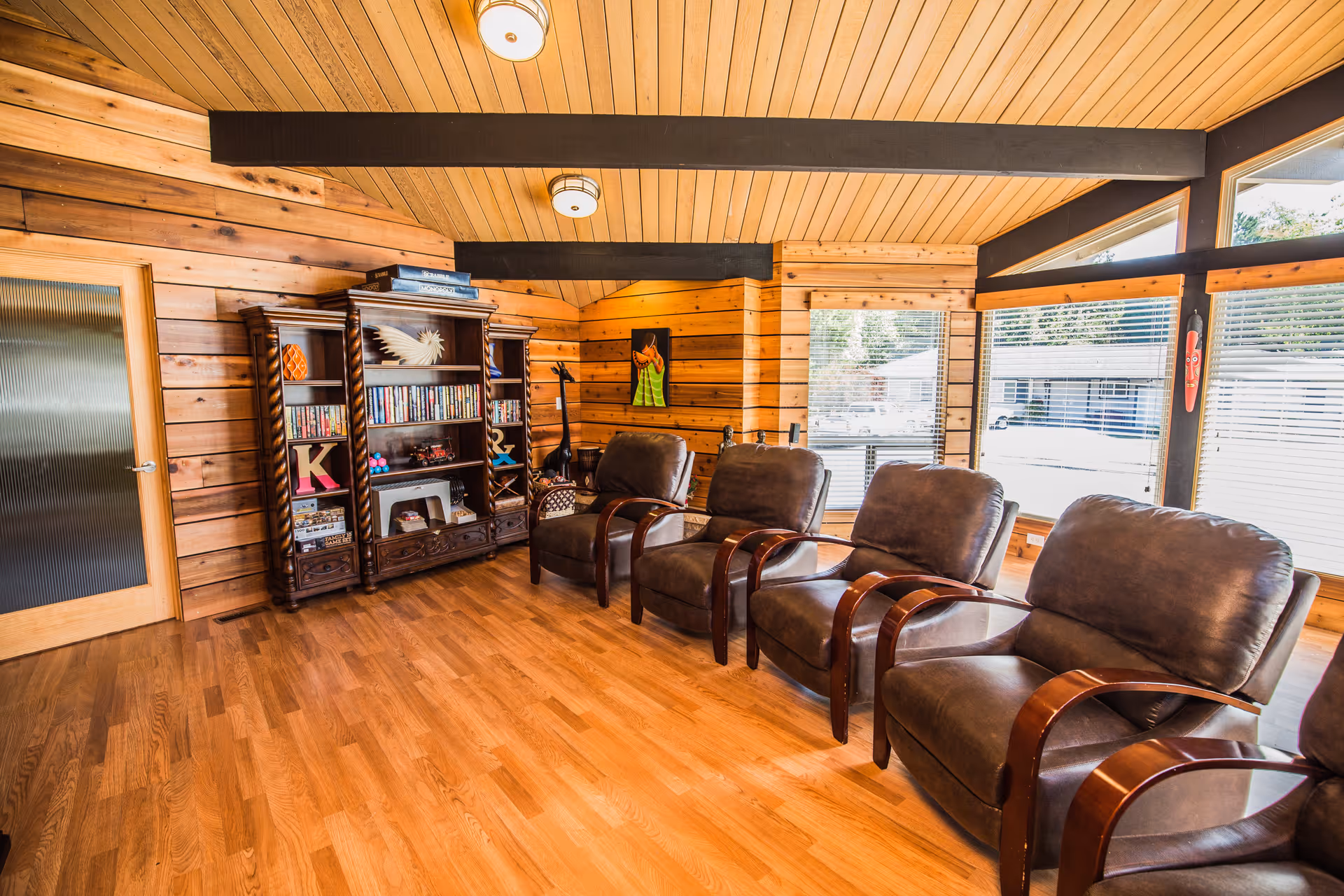 A cozy living room with wooden walls and ceiling, featuring four brown cushioned armchairs with wooden armrests arranged in a row. There is a wooden bookshelf filled with books and decorative items against the wall. Large windows with blinds allow natural light to fill the room, and a wooden door with frosted glass is visible on the left side.