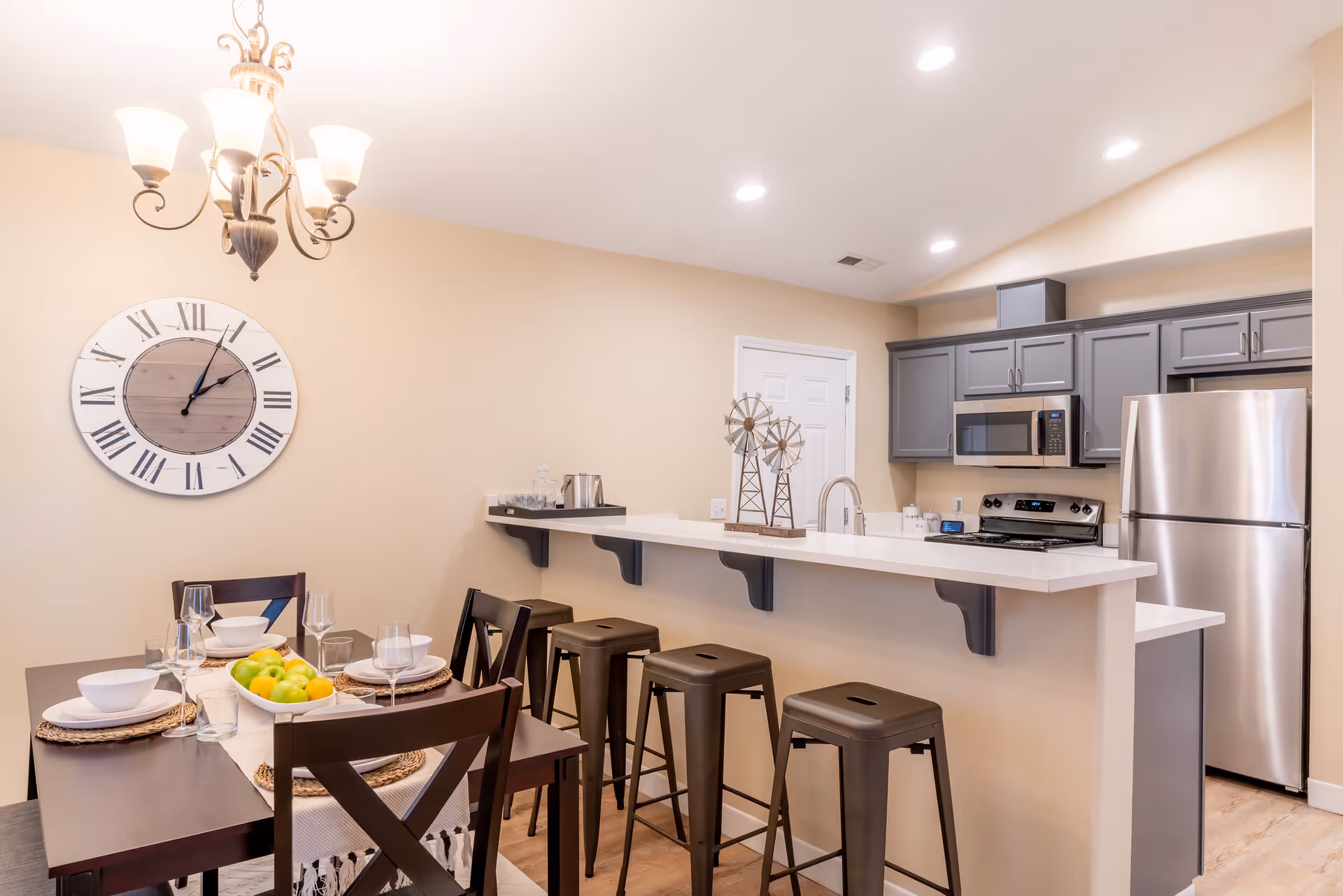 A modern kitchen and dining area in a senior living facility. The kitchen features gray cabinets, stainless steel appliances including a refrigerator, microwave, and stove. A white countertop bar with four black bar stools separates the kitchen from the dining area. The dining table is set with white bowls, glasses, and a centerpiece of green and yellow apples. A large wall clock with Roman numerals hangs on the beige wall, and a chandelier with five lights is above the dining table.