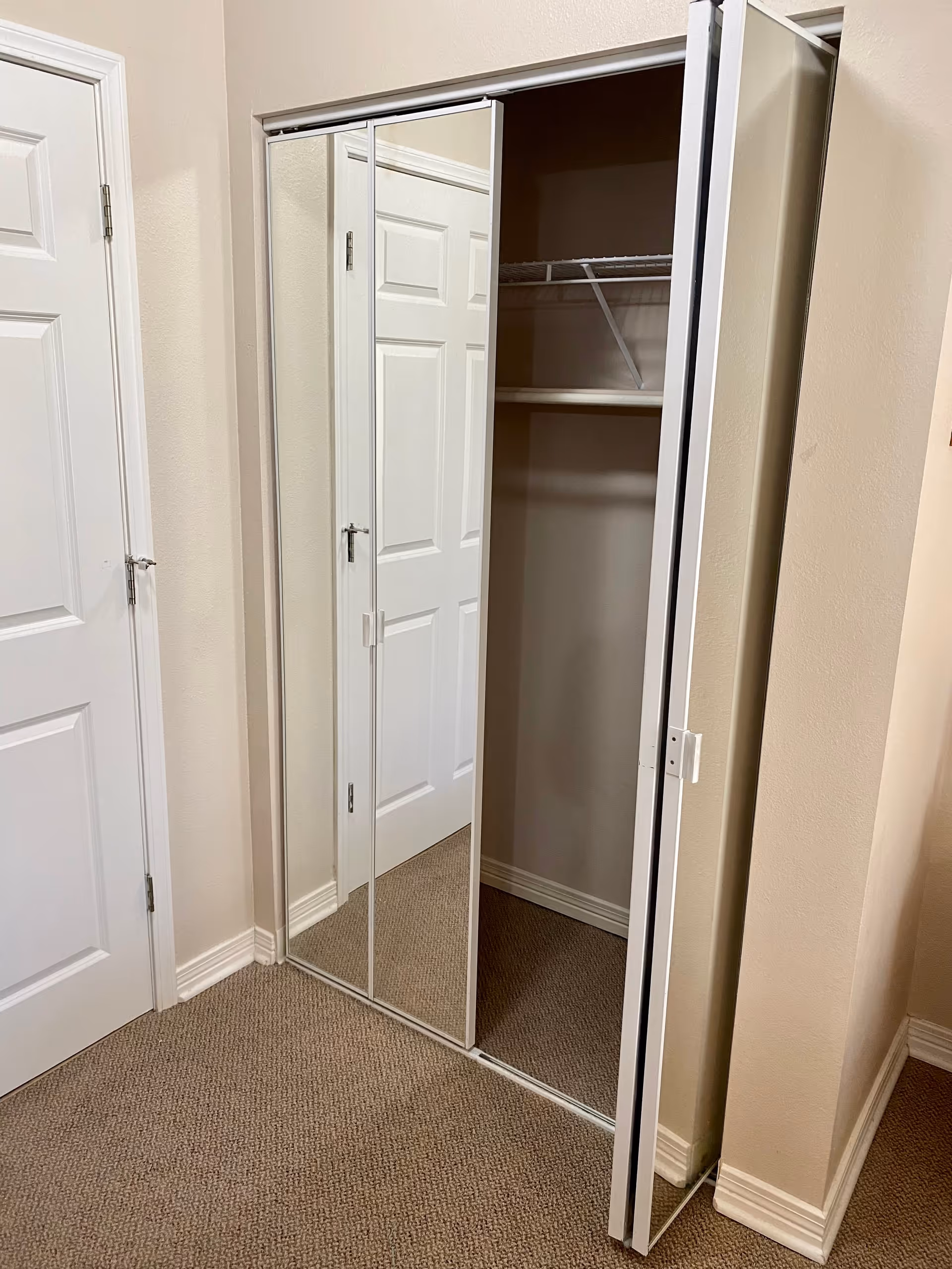An empty closet with sliding mirrored doors partially open, showing a shelf and hanging rod inside. The closet is in a room with beige walls and carpeted floor, next to a closed white door.