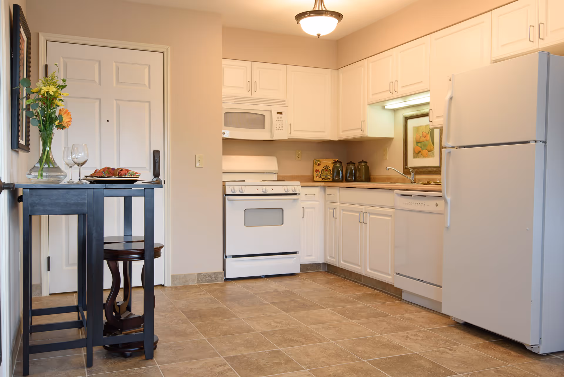A clean kitchen with white cabinets, a white refrigerator, stove, microwave, and dishwasher. There is a small black table with two wine glasses, a plate with napkins, and a vase with flowers near a white door. The floor is tiled and the walls are painted beige.