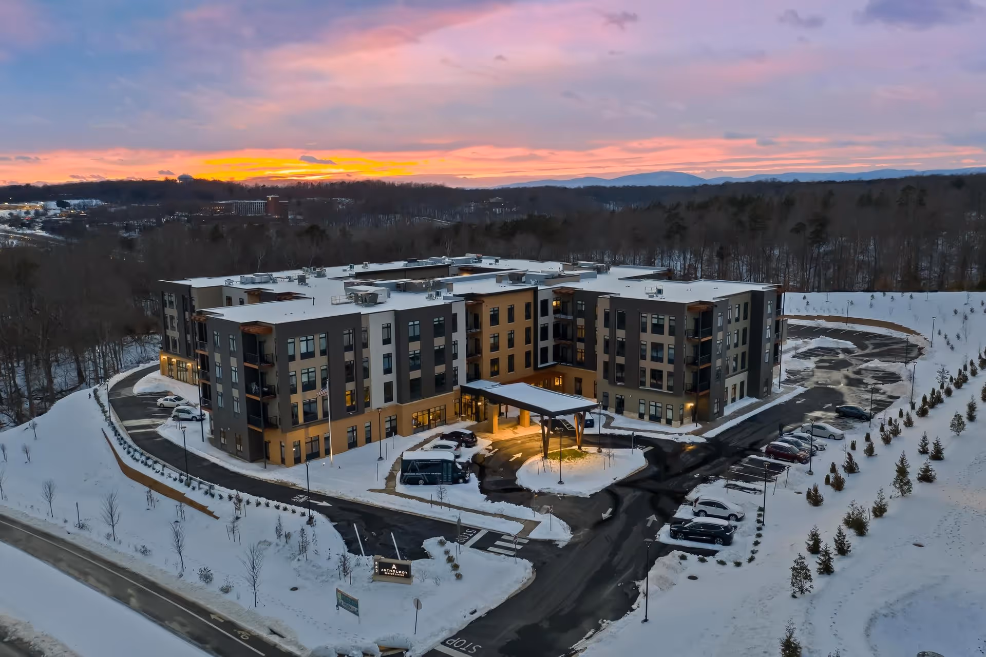 Aerial view of The Barclay at Charlottesville senior living facility during winter at sunset, showing a large modern four-story building surrounded by snow-covered grounds and parking areas with several cars parked.