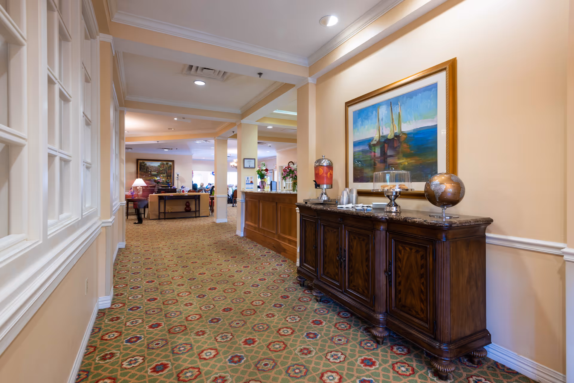 Carpeted interior hallway with a wooden sideboard holding a globe and beverage dispenser and a framed painting, leading into a lounge area.