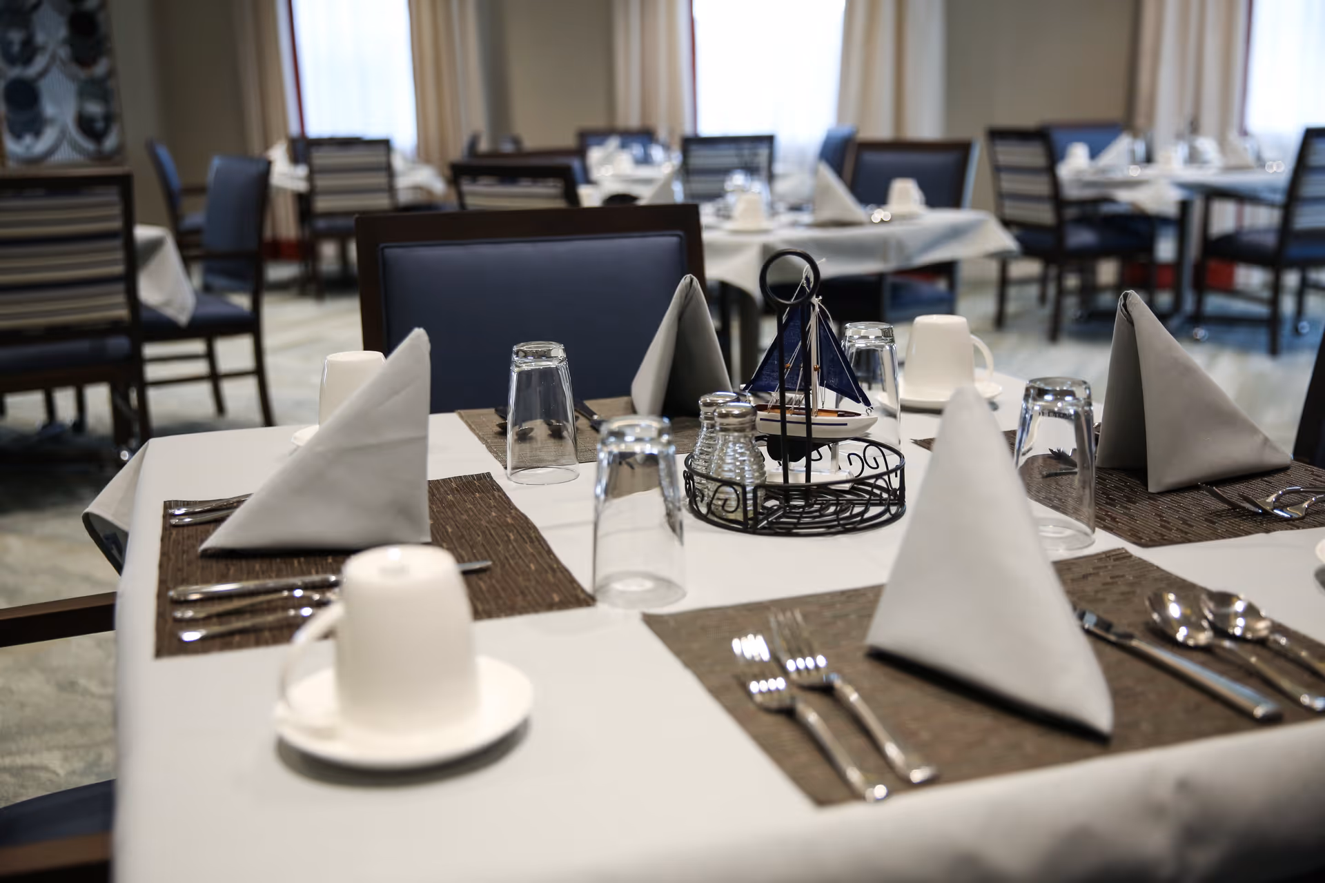 A dining table set for a meal with folded gray napkins, upside-down drinking glasses, white coffee cups on saucers, silverware, and a nautical-themed centerpiece featuring a small sailboat. Several other similarly set tables and chairs are visible in the background in a well-lit dining room.