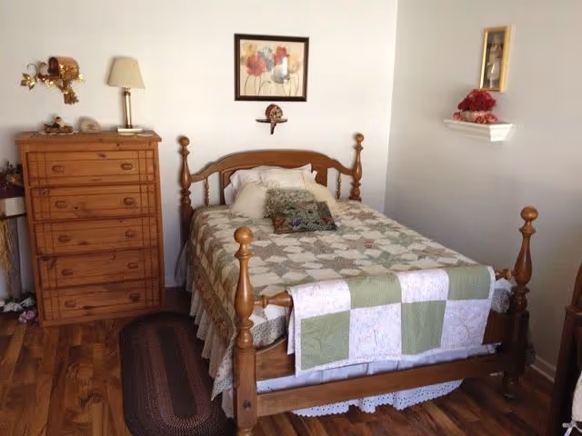 A cozy bedroom with a wooden bed frame featuring a quilted bedspread and decorative pillows. To the left of the bed is a wooden dresser with a lamp and small decorative items on top. The walls are light-colored with a framed floral picture above the bed and a small shelf with a flower arrangement on the right wall. The floor is wooden with a small rug beside the bed.