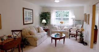 A cozy living room with a beige sofa, two armchairs, a round wooden coffee table with decorative items, a side table with a lamp, a large window with white curtains, framed artwork on the walls, and a potted plant in the corner.