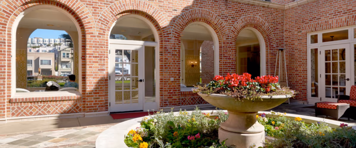 Outdoor courtyard area with a large stone planter filled with red and yellow flowers in the center. The courtyard is surrounded by a brick building with arched windows and glass doors. There is outdoor seating with red cushions visible on the right side.