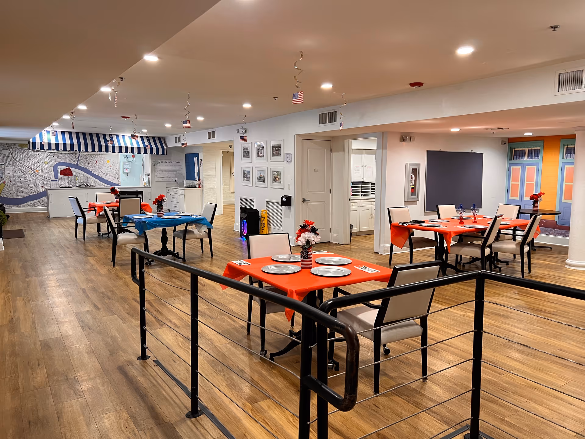 Interior view of a dining area in an assisted living facility with several tables covered in red and blue tablecloths, each set with plates, utensils, and small flower centerpieces. The room has wood flooring, white walls, and ceiling lights. A large map mural is visible on the far wall, and there is a railing in the foreground.