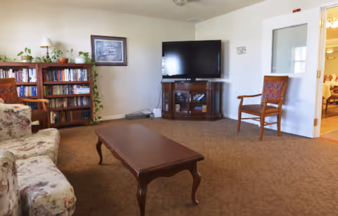 A cozy living room with a floral patterned sofa, a wooden coffee table, a wooden bookshelf filled with books and plants on top, a flat-screen TV on a wooden TV stand, and a wooden chair with a patterned cushion. The room has beige walls and carpet, and a door leading to another room is visible.