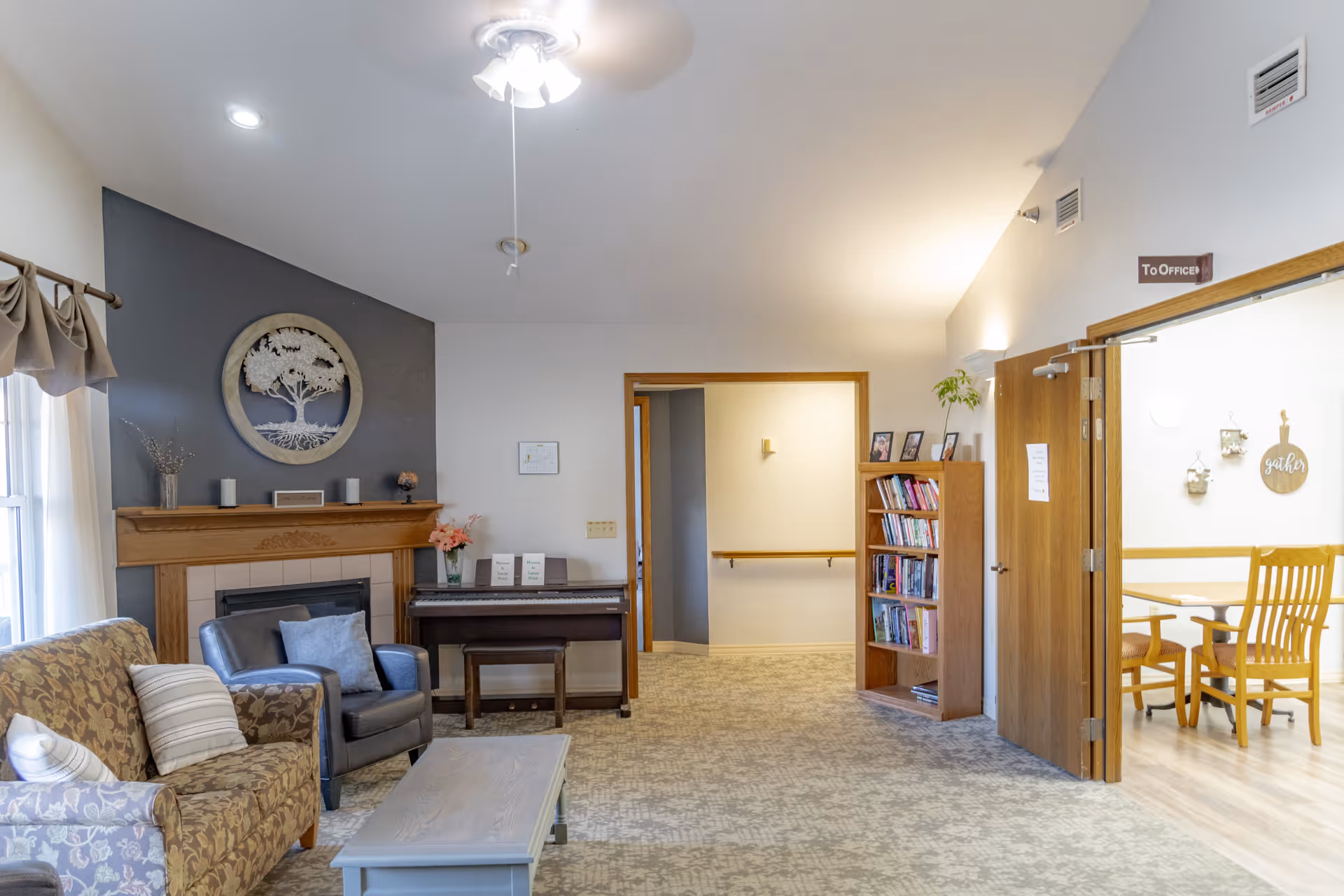 A cozy living room area with a patterned sofa, a gray armchair, and a light blue coffee table. There is a wooden fireplace with a decorative tree wall art above it, and a piano with a bench next to the fireplace. A bookshelf filled with books is positioned near an open doorway leading to a dining area with wooden chairs and a table. The room is well-lit with natural light coming through a window with curtains and a ceiling fan with lights.
