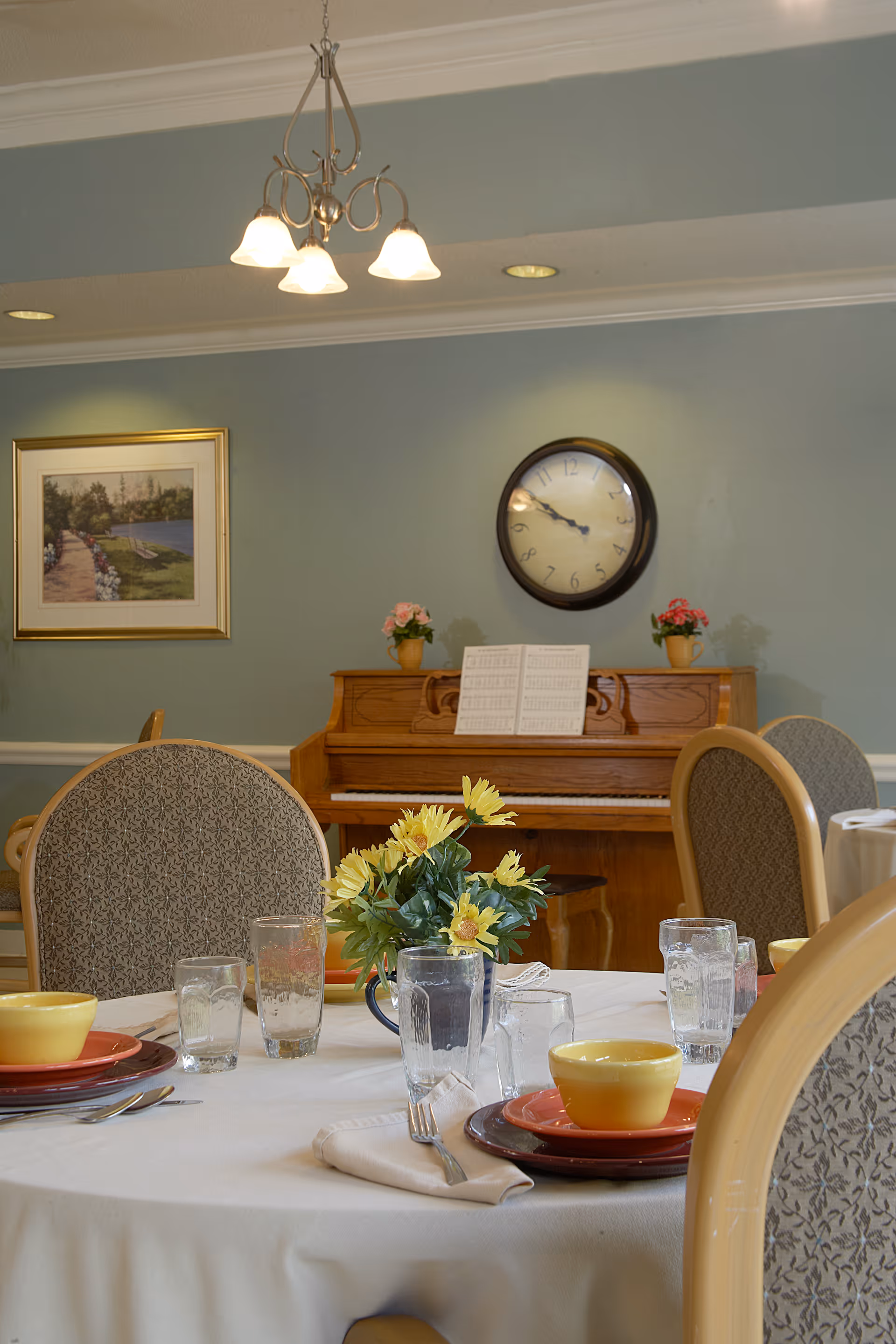 A dining room table set with yellow bowls, red plates, glasses, and silverware on a white tablecloth. In the background, there is a wooden piano with sheet music and two small flower pots on top. A round wall clock and a framed picture hang on the light blue wall. Upholstered dining chairs surround the table.