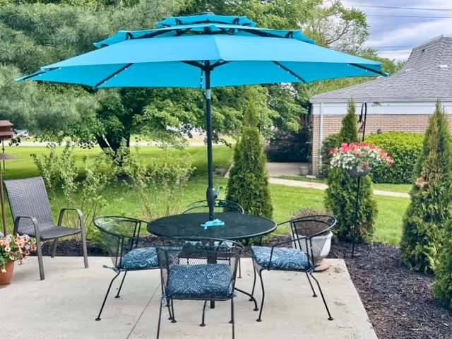 Outdoor patio area with a round black metal table and four matching chairs with blue cushions, shaded by a large blue umbrella. Surrounding the patio are green shrubs, small trees, and a hanging flower basket with pink and white flowers. A brick building and a grassy lawn are visible in the background.