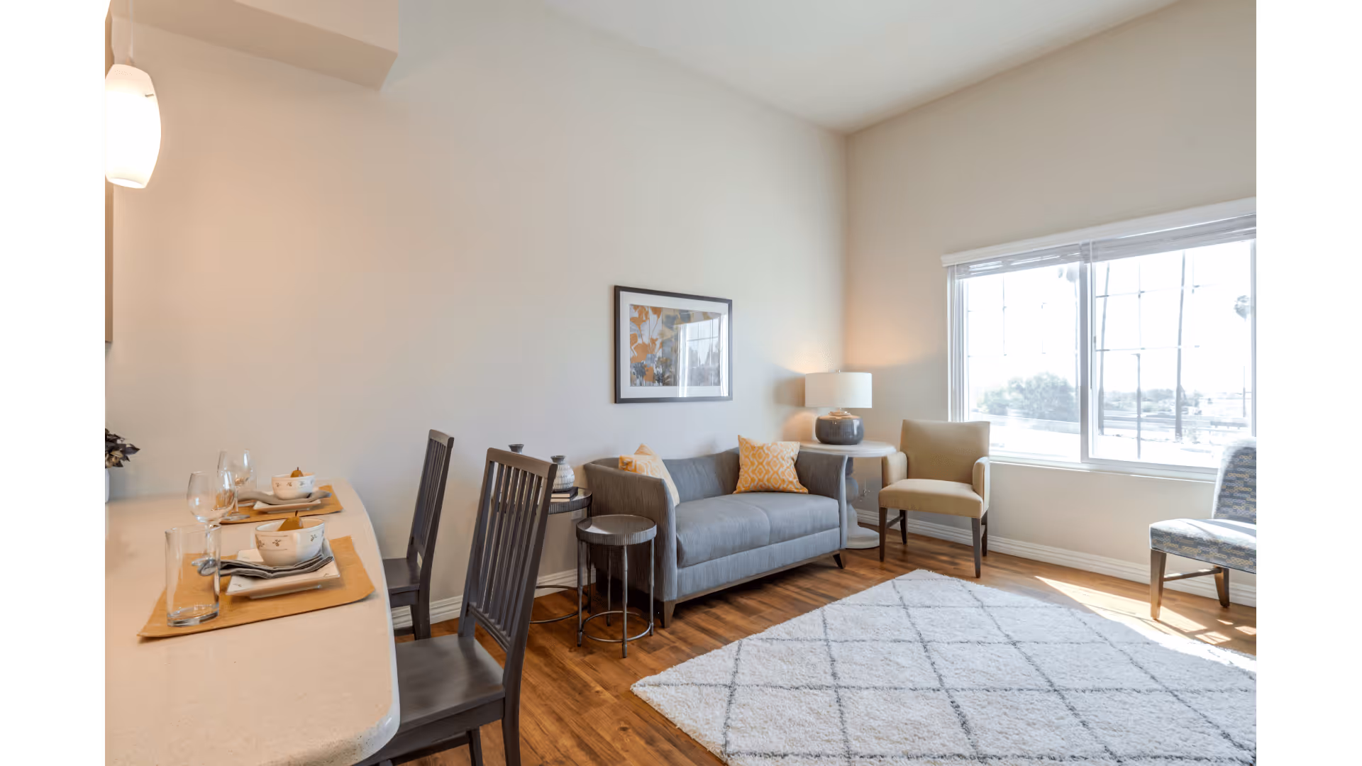 A bright living room with a large window letting in natural light. The room features a gray sofa with yellow and gray cushions, a side table with a lamp, two chairs, and a white rug with a diamond pattern on a wooden floor. To the left, there is a countertop with place settings including plates, bowls, glasses, and napkins.