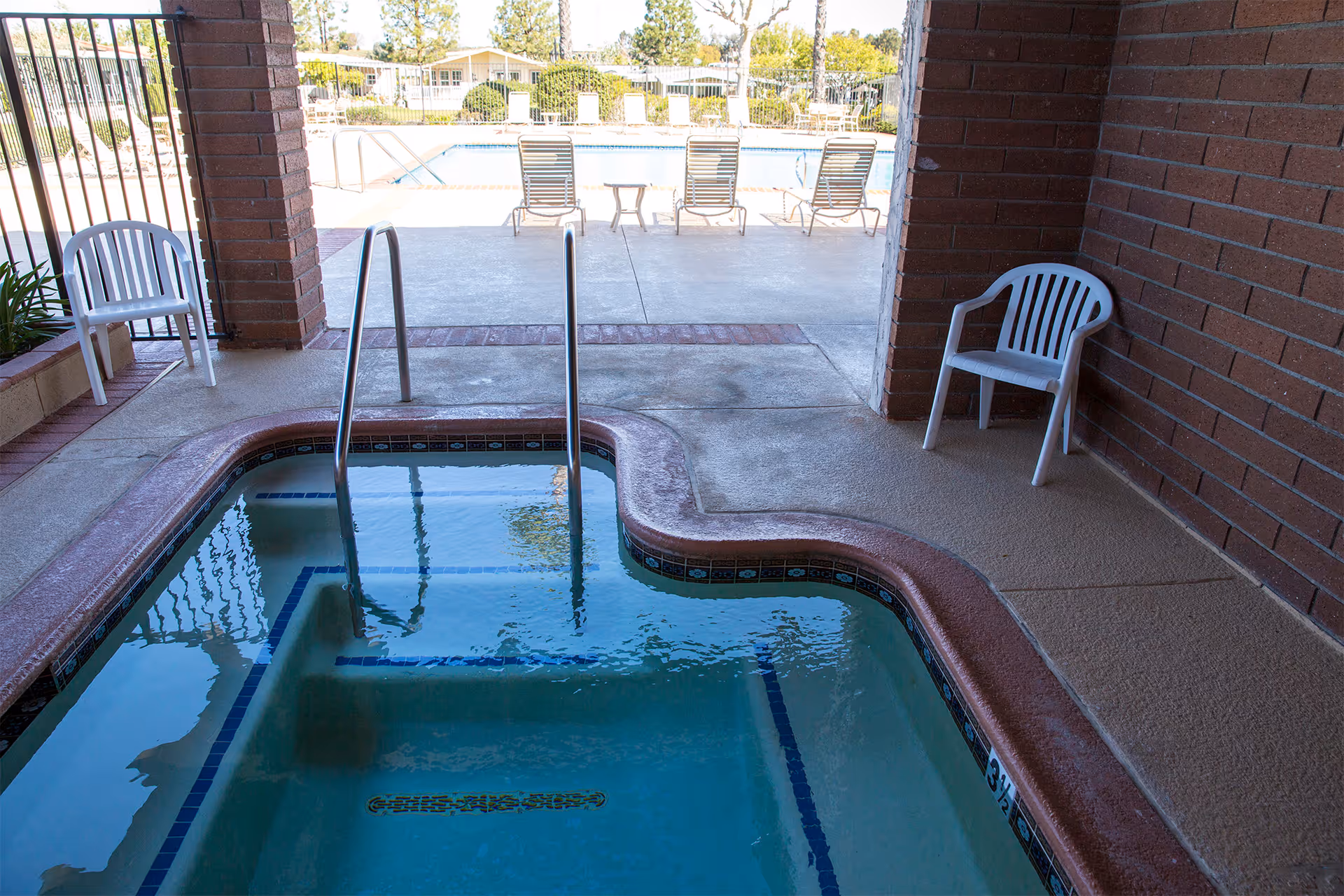 Indoor hot tub with metal handrails and tiled edges, two white plastic chairs on the surrounding concrete floor, and an outdoor swimming pool with lounge chairs visible through an open area framed by brick walls.