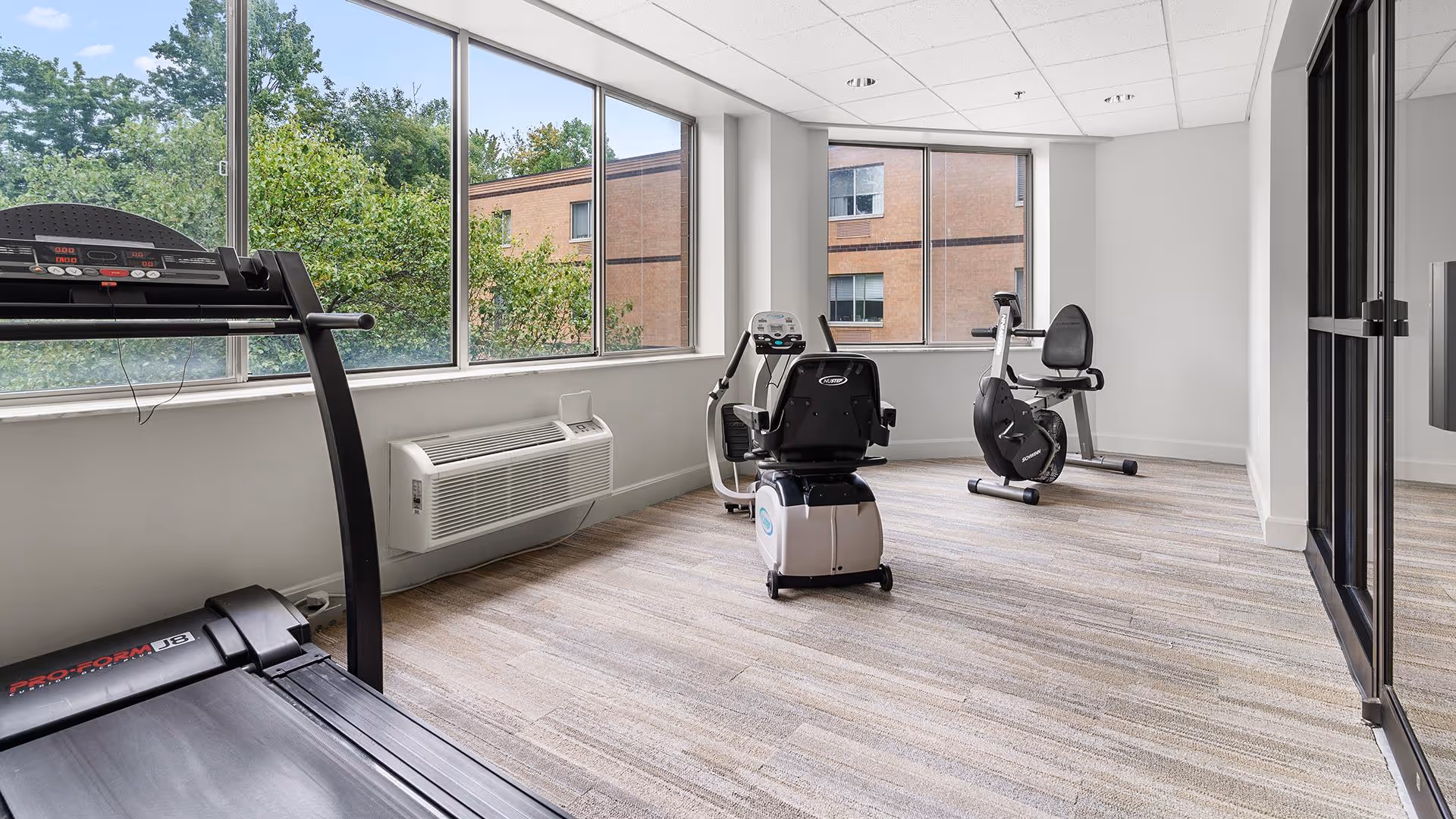 A bright exercise room with large windows showing greenery and a building outside. Inside, there is a treadmill, a recumbent exercise bike, and an upright exercise bike on a light-colored wood floor. The walls are white, and there is a wall-mounted air conditioning unit beneath the windows.