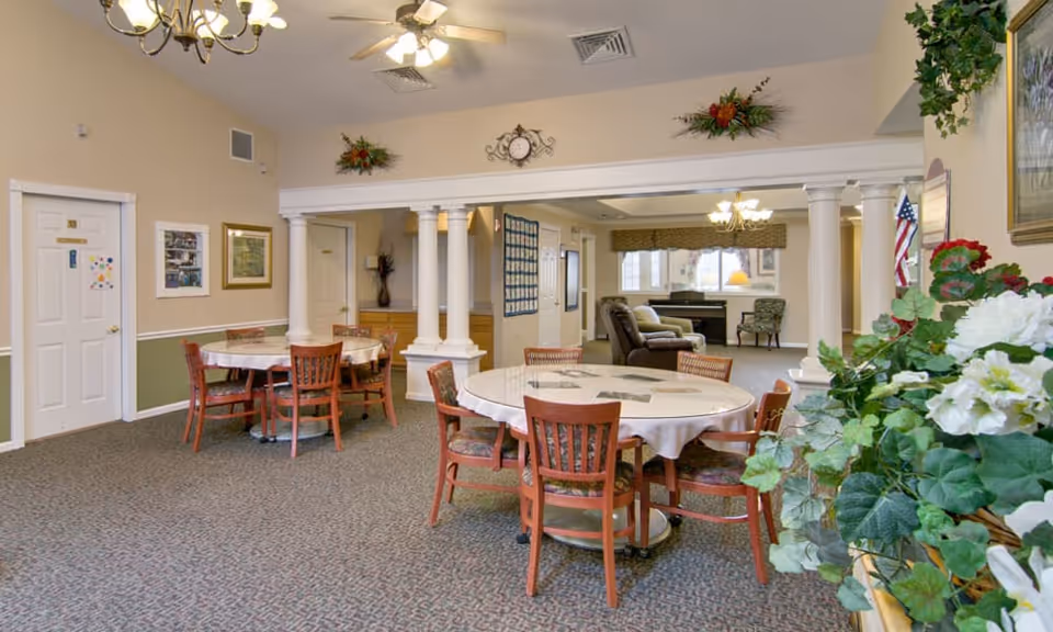 A spacious common area in a senior living facility with round tables covered with white tablecloths and surrounded by wooden chairs. The room features beige walls with white trim, decorative columns, ceiling fans with lights, and floral arrangements. In the background, there is a seating area with armchairs and a piano near windows with valances.