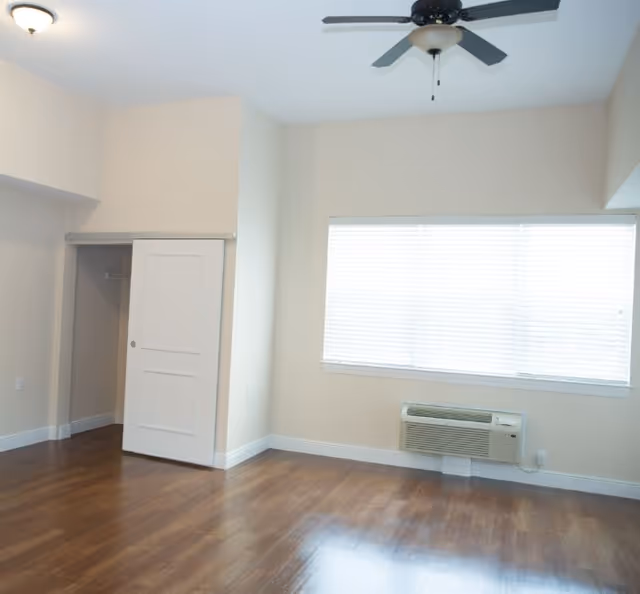 Empty room with wooden flooring, a ceiling fan, a window with blinds, a wall-mounted air conditioning unit, and an open closet with a sliding door.