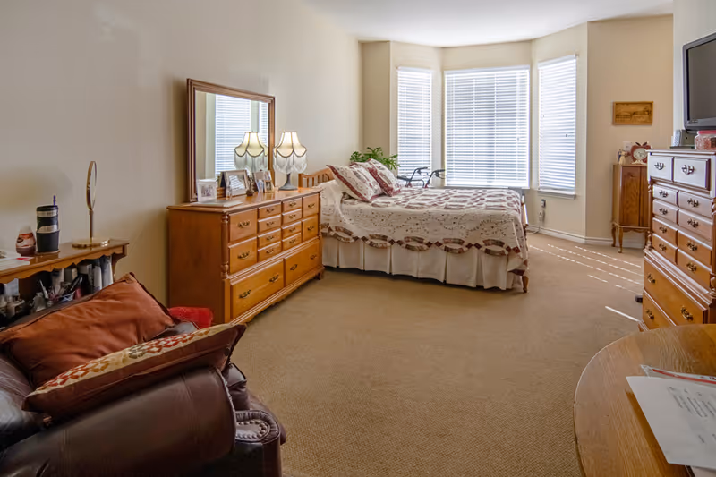 A well-lit bedroom with a large bed covered in a patterned quilt, two wooden dressers with lamps and framed photos, a leather armchair with cushions, and windows with white blinds letting in natural light.