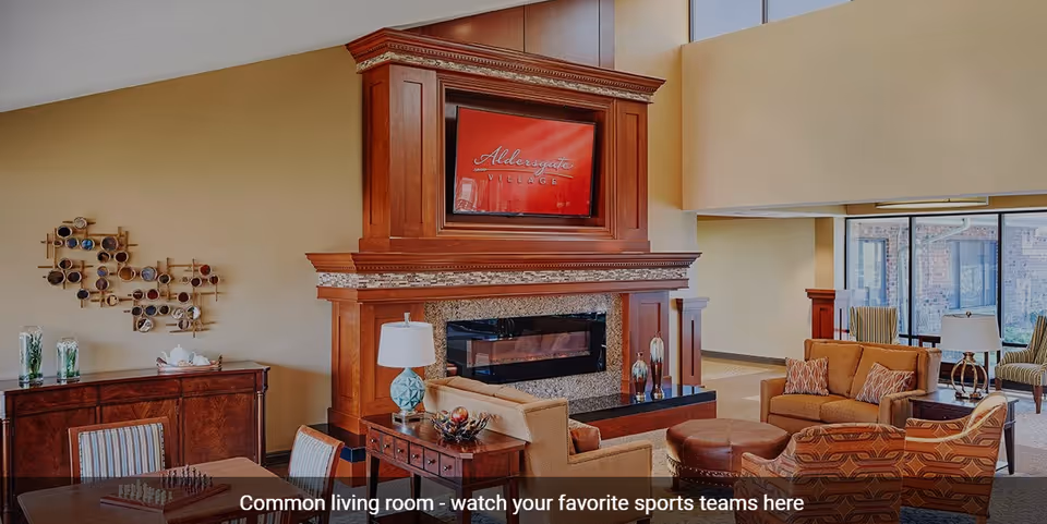 A spacious common living room with comfortable seating including armchairs and sofas arranged around a large wooden fireplace with a mounted TV displaying 'Aldersgate Village'. The room features warm lighting, a decorative wall piece, and large windows allowing natural light to enter.