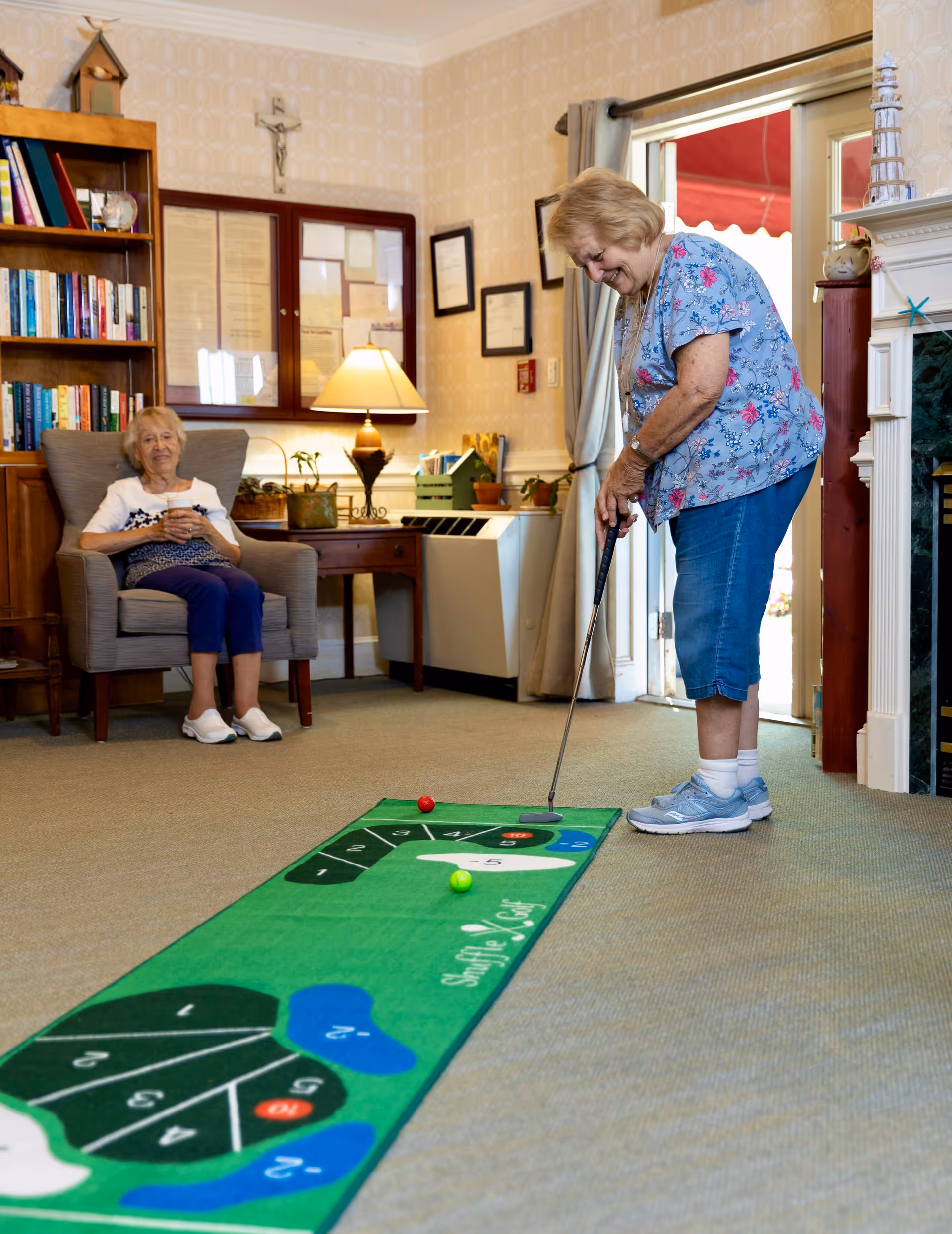 An elderly woman playing indoor shuffleboard golf on a green mat in a cozy living room. Another elderly woman is sitting comfortably in a chair nearby, watching and smiling. The room has a bookshelf, a lamp on a wooden table, framed pictures on the wall, and a fireplace.