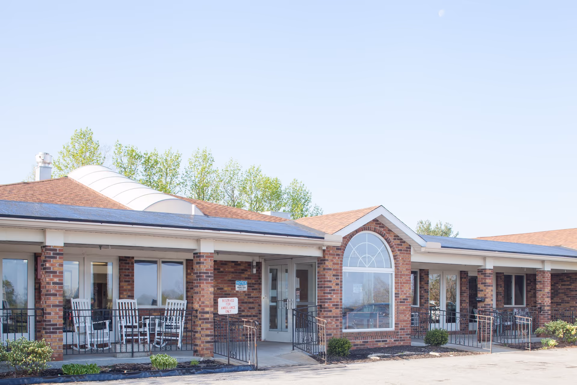 Front exterior of a single-story brick healthcare building with a covered porch, rocking chairs, and an arched window.