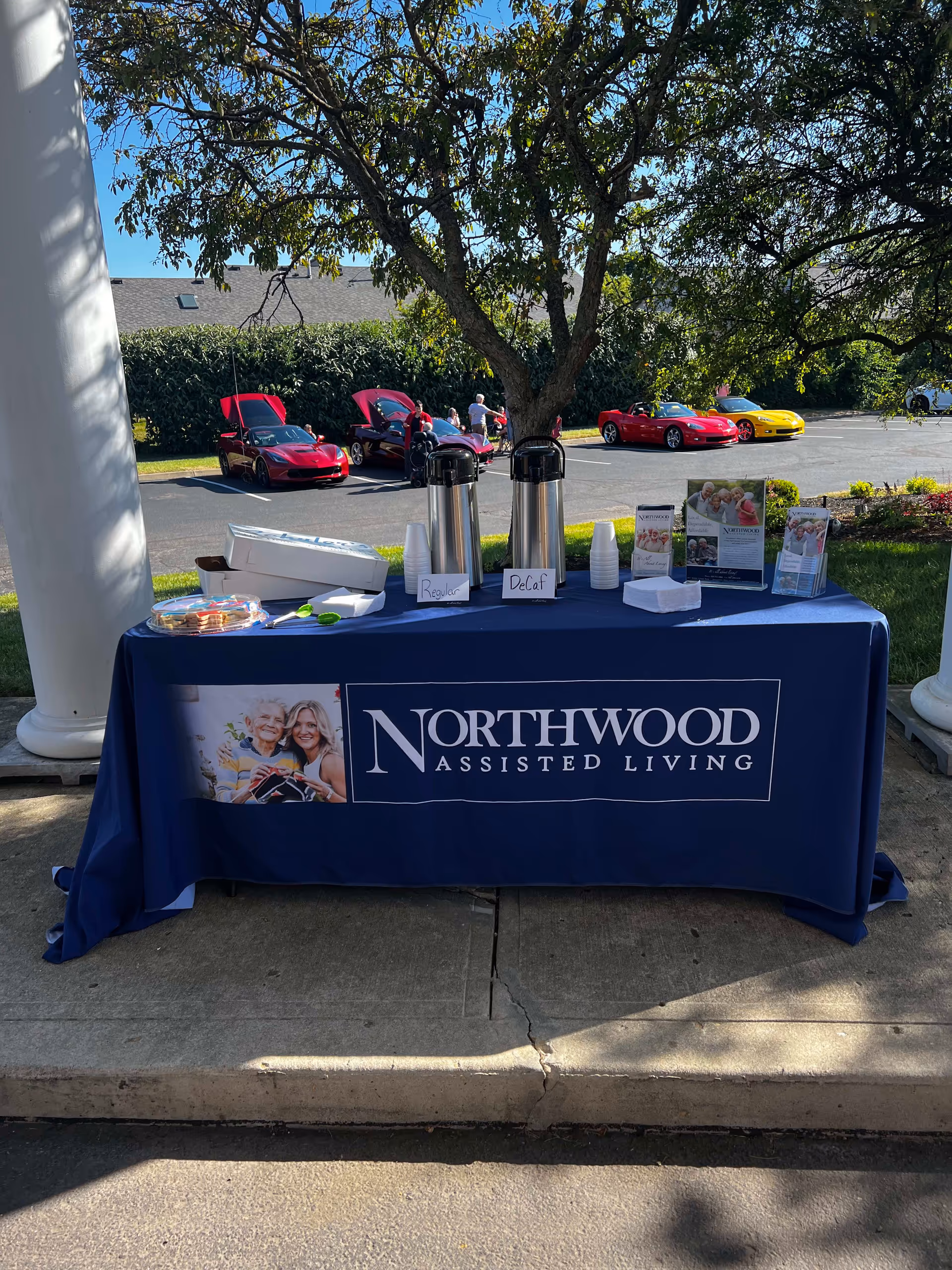 Outdoor table covered with a blue tablecloth displaying the Northwood Assisted Living logo. On the table are two coffee dispensers labeled Regular and Decaf, stacks of cups, napkins, brochures, and a plate of cookies. In the background, there are several red and yellow sports cars with their hoods open, people standing around, and a tree providing shade.