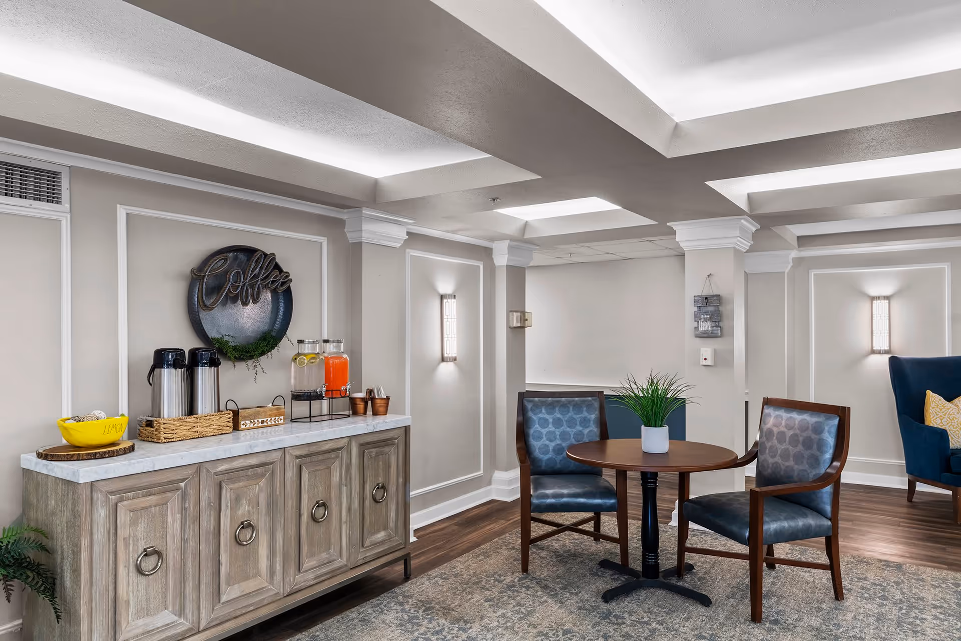 A cozy seating area in a senior living facility with a small round wooden table and two blue cushioned chairs. To the left, there is a wooden sideboard with a marble top holding coffee dispensers, a yellow bowl labeled 'Lemon', and two beverage dispensers with orange and lemon-infused water. The wall behind the sideboard has a decorative round sign that says 'Coffee'. The room features soft lighting, white crown molding, and a patterned area rug on wooden flooring.