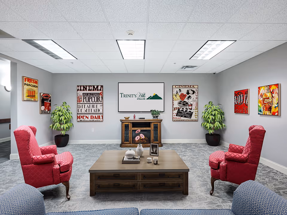 A cozy lounge area with two red upholstered armchairs facing a wooden coffee table with decorative items. The room has gray walls adorned with vintage cinema-themed posters and two large potted plants. A wooden cabinet with a framed sign reading 'Trinity Hills of Knoxville' is centered on the back wall under a white ceiling with fluorescent lights.