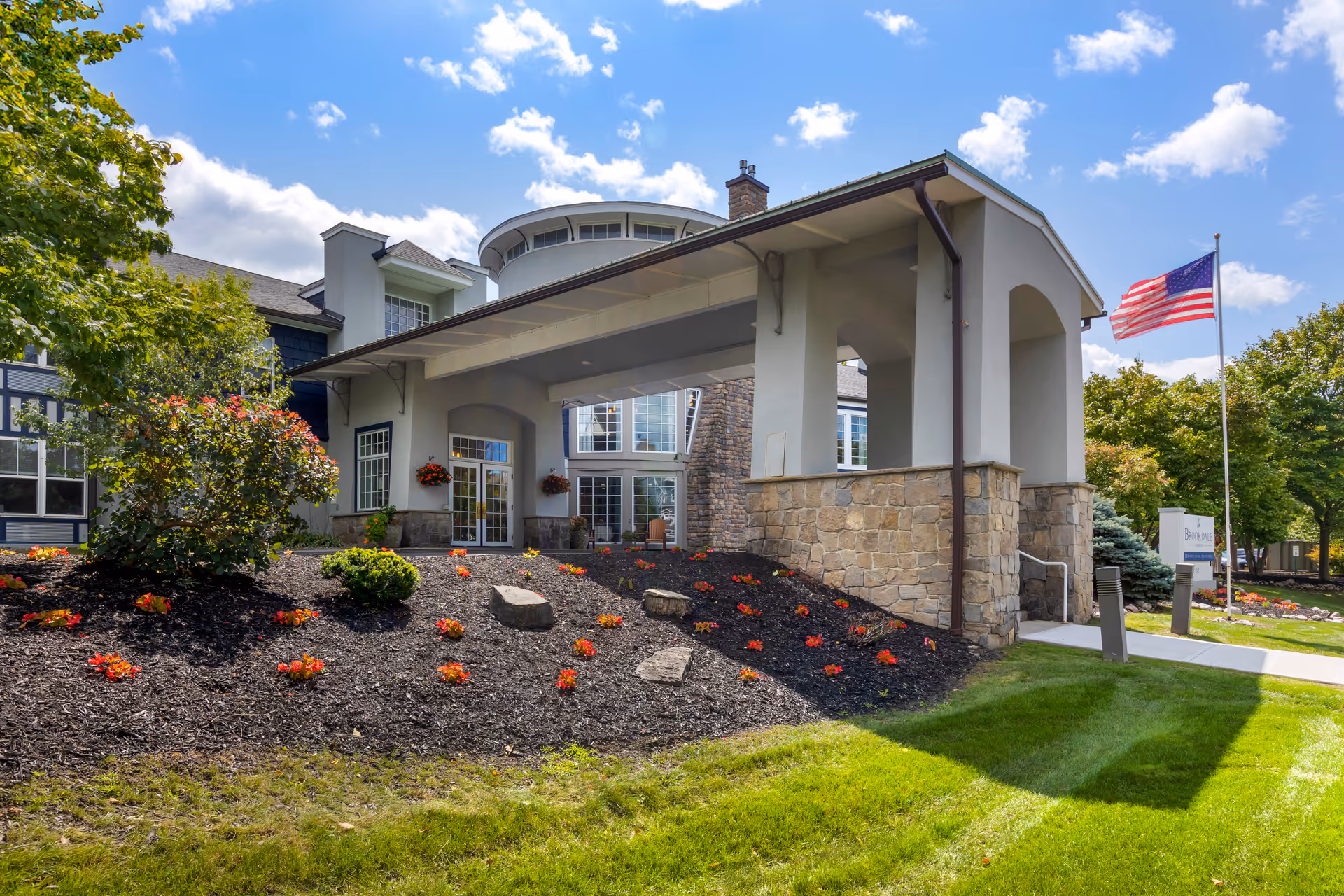 Exterior view of the Brookdale Manlius senior living facility entrance with a covered drop-off area, stone and stucco facade, landscaped flower beds with red flowers, green lawn, trees, and an American flag on a flagpole under a partly cloudy blue sky.