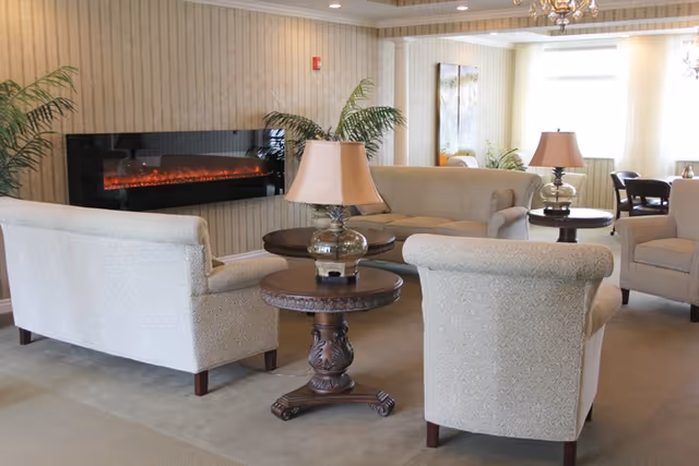 A cozy living room area with beige upholstered sofas and armchairs arranged around wooden side tables with lamps. A modern electric fireplace is mounted on the wall, and there are green plants and framed artwork in the background. The room has soft lighting and a carpeted floor.