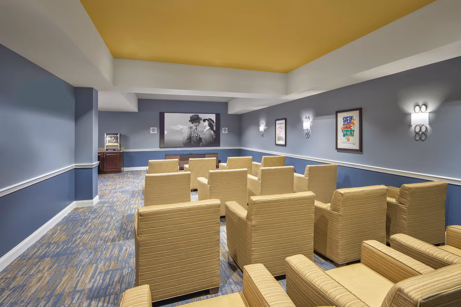A small movie theater room with multiple rows of beige armchairs facing a large screen showing a black and white movie. The room has blue walls with white trim, a yellow ceiling, wall sconces, framed movie posters, and a popcorn machine in the back corner.