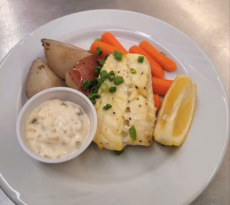 A plated meal of baked white fish topped with chopped green onions, served with roasted potatoes, baby carrots, a lemon wedge, and a cup of tartar sauce.