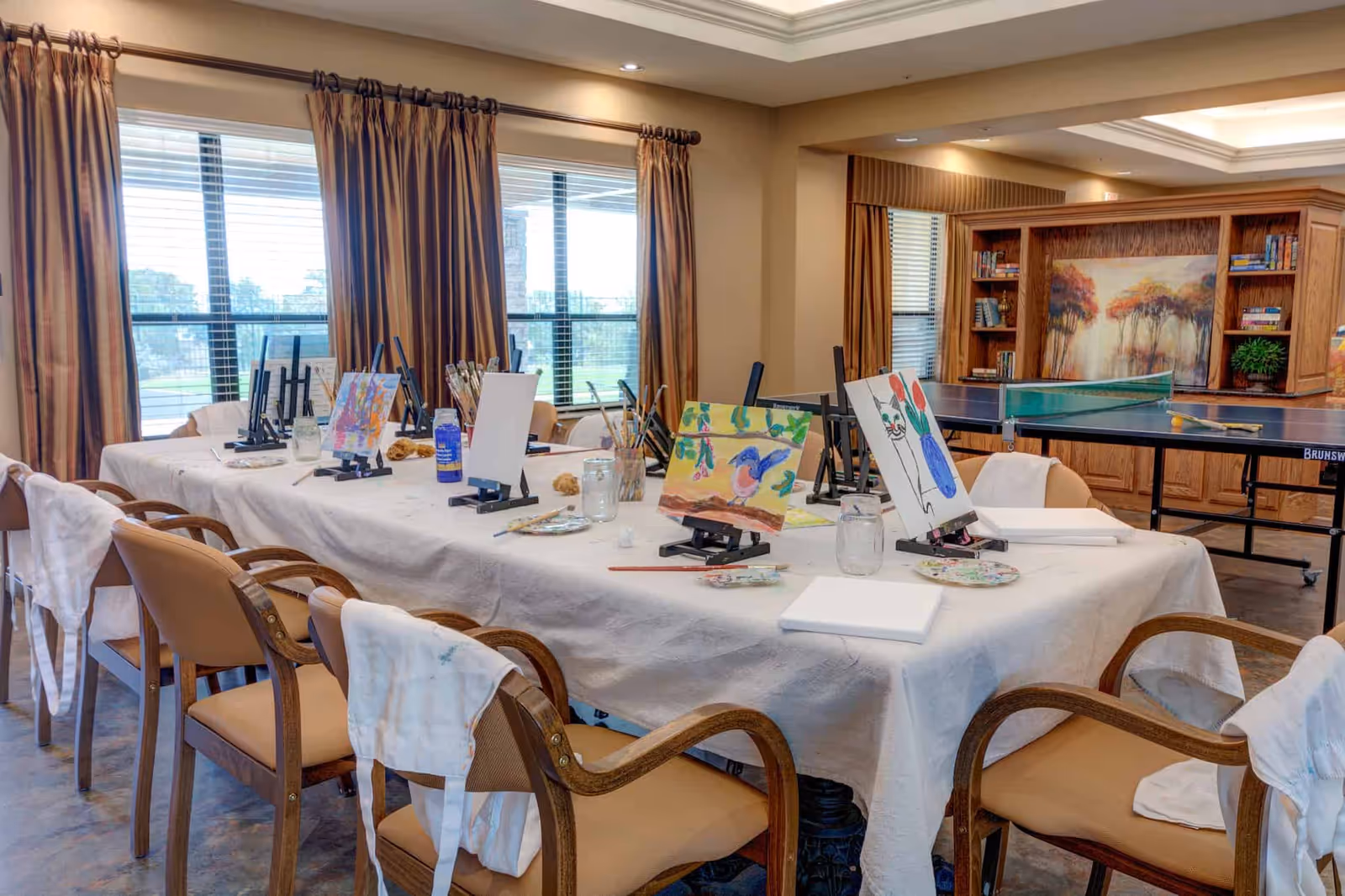 A bright activity room with a long table set for painting—easels, canvases, brushes and aprons on chairs—with a ping-pong table and bookshelves in the background.