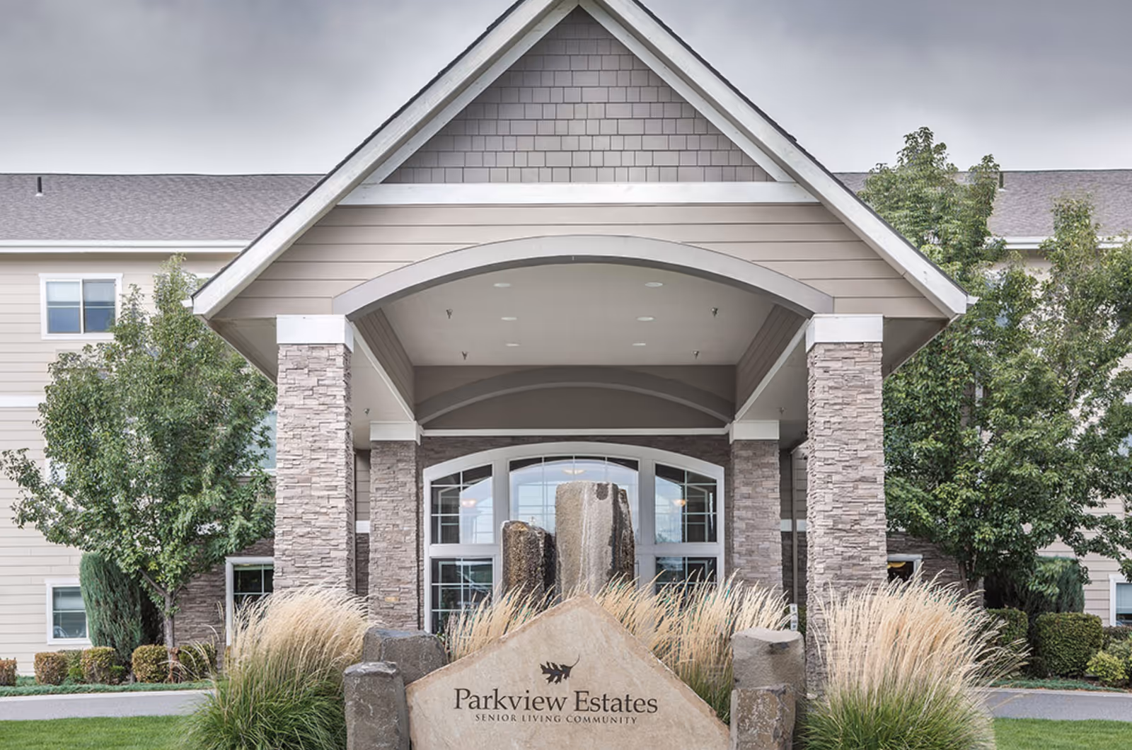 Front entrance of Parkview Estates senior living community featuring a covered driveway supported by stone pillars, landscaped with ornamental grasses and trees, and a large stone sign with the facility's name.