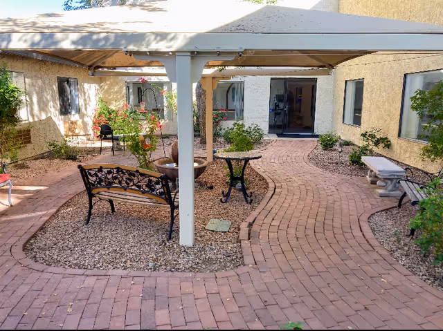 Outdoor courtyard area with a covered patio, brick pathways, benches, small tables, and various plants and shrubs surrounding the space. The courtyard is enclosed by a beige building with several windows and a glass door.