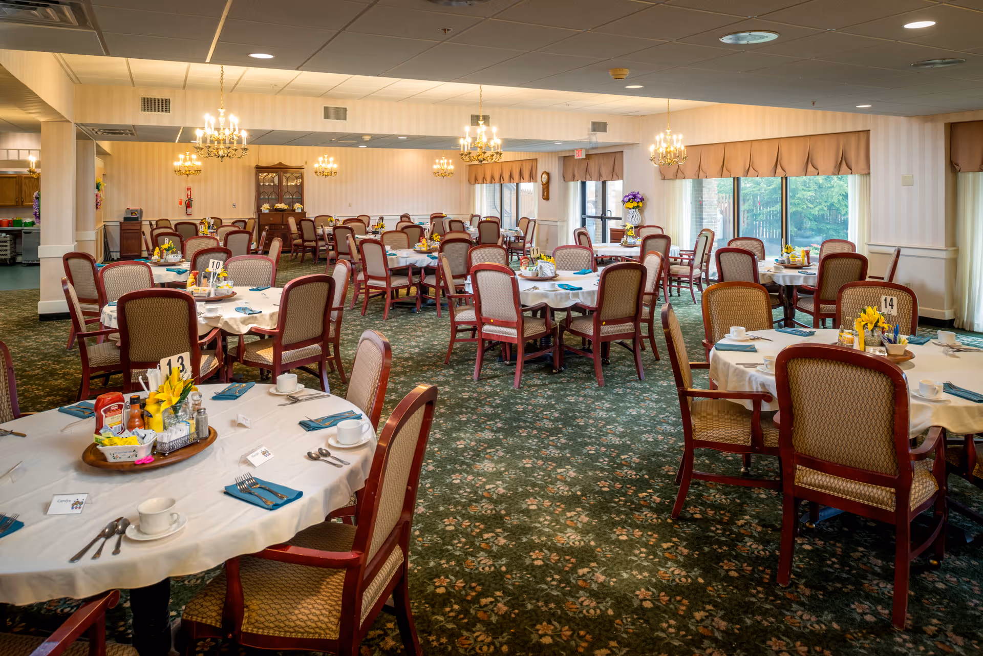 A spacious dining room with multiple round tables covered with white tablecloths and set with cups, saucers, utensils, and blue napkins. Each table has a centerpiece with condiments and flowers. The room has green floral carpet, beige walls with subtle stripes, large windows with brown valances, and several chandeliers hanging from the ceiling providing warm lighting.