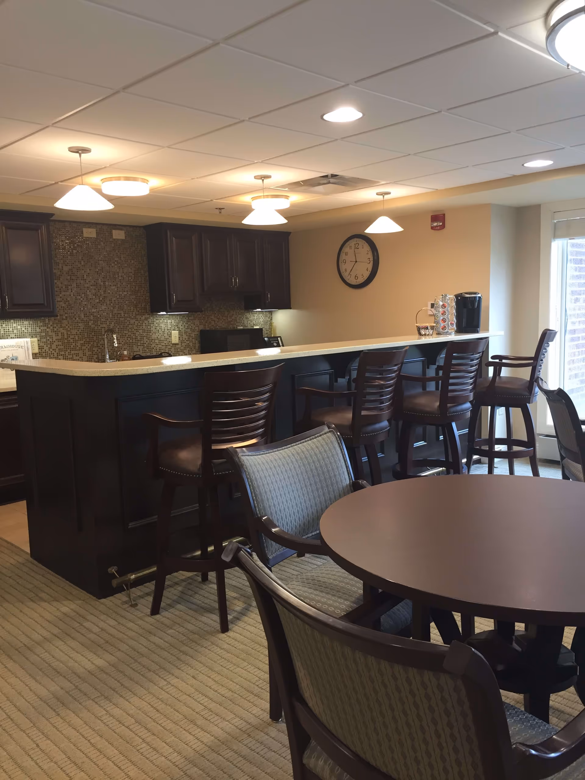 Interior view of a communal dining and kitchen area with a round table and chairs in the foreground, a kitchen counter with bar stools, dark wood cabinetry, a mosaic tile backsplash, and a wall clock showing the time as 6:15.