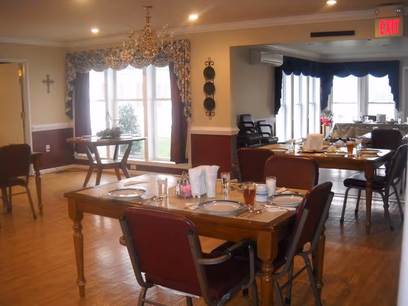 A dining room with wooden tables set for meals, upholstered chairs, a chandelier, and large windows.