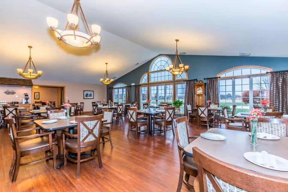 A spacious dining room with multiple wooden tables and chairs arranged neatly. Each table is set with plates, napkins, and vases containing flowers. Large windows with arched tops allow natural light to fill the room, and elegant chandeliers hang from the ceiling. The floor is wooden, and there is a grandfather clock near the windows.
